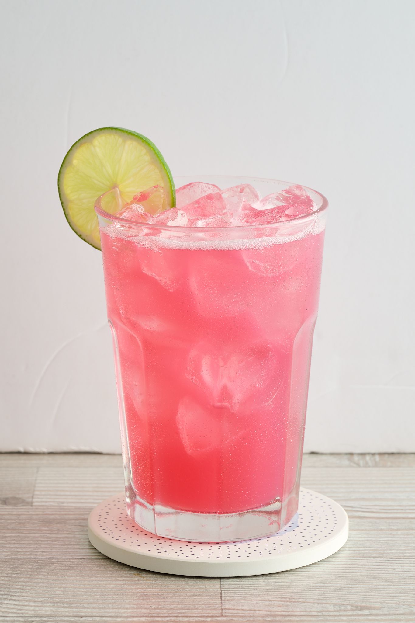 Pink iced drink with a lime slice in a clear glass on a white saucer