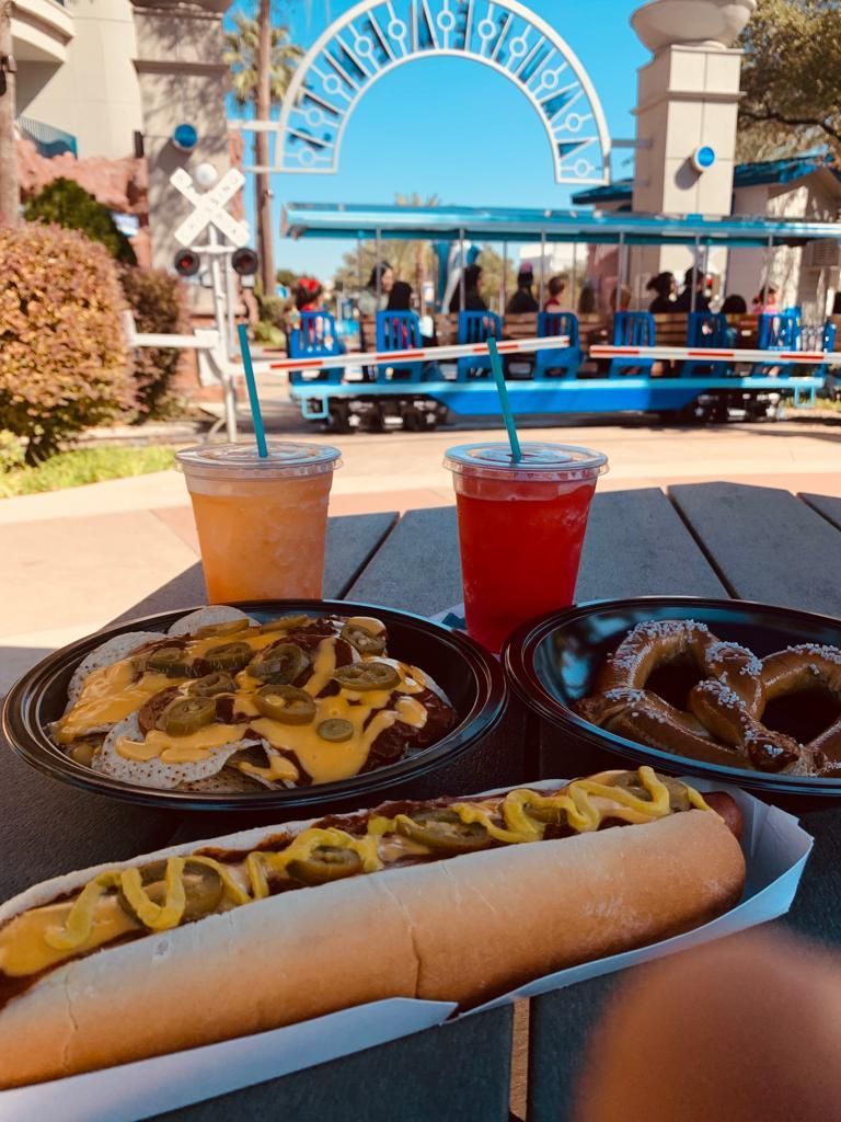 A hot dog a pretzel and a drink on a table with a train in the background
