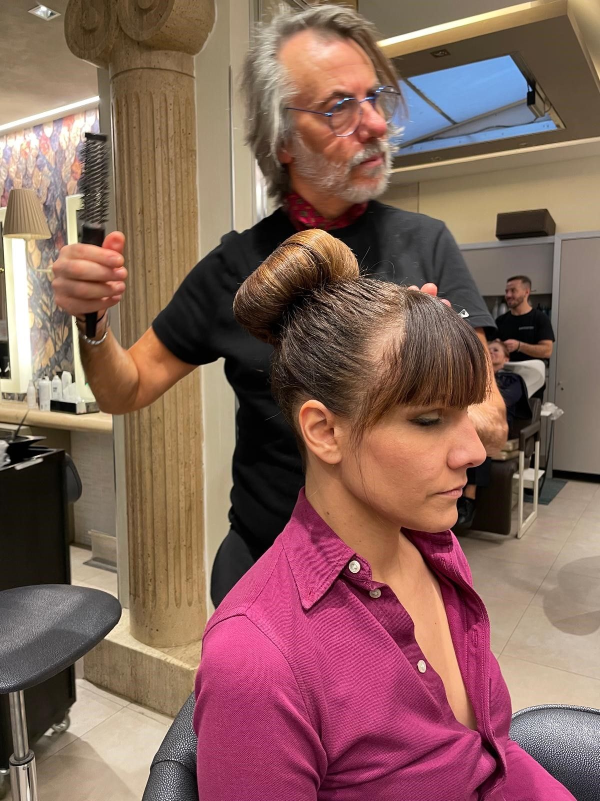 In un salone, una parrucchiera pettina i capelli di una cliente, che è seduta con un grande chignon ben acconciato in testa.