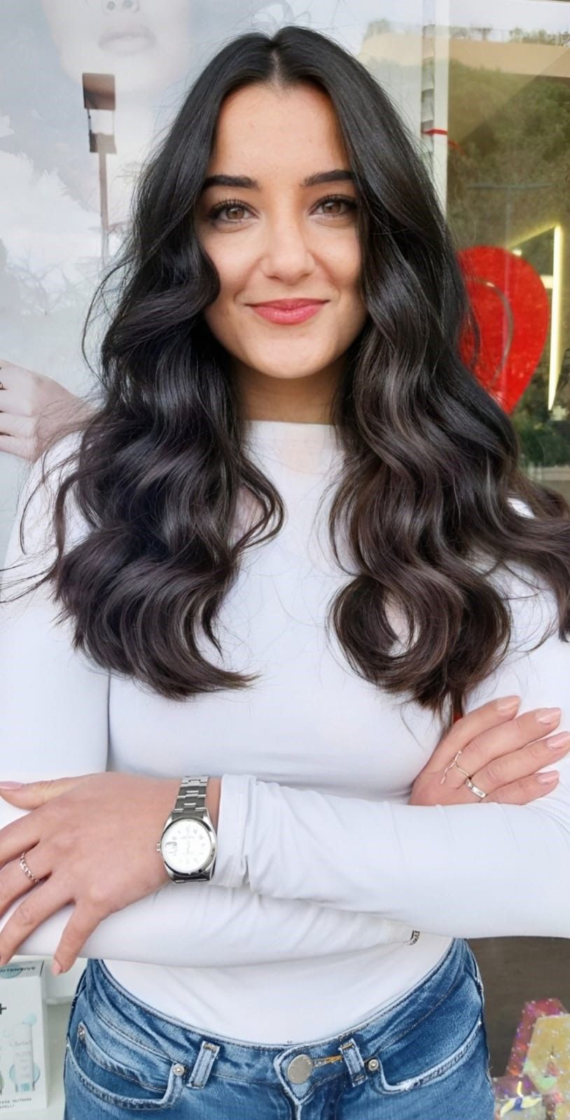 Una persona sorridente con lunghi capelli scuri e ondulati, che indossa una camicia bianca a maniche lunghe e un orologio, con le braccia incrociate in una posa informale.