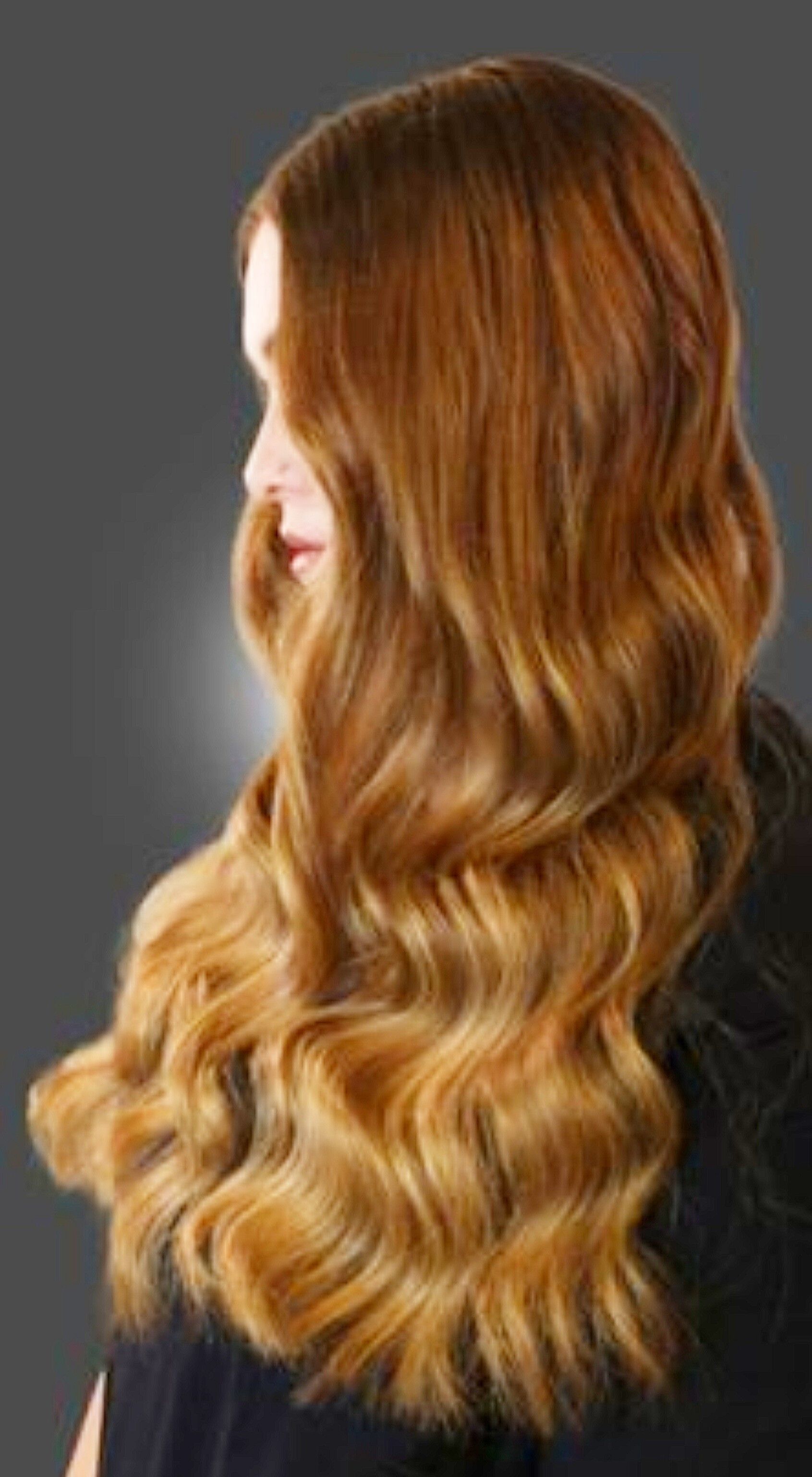 Una vista di profilo di lunghi capelli ondulati color castano dorato su uno sfondo grigio uniforme.