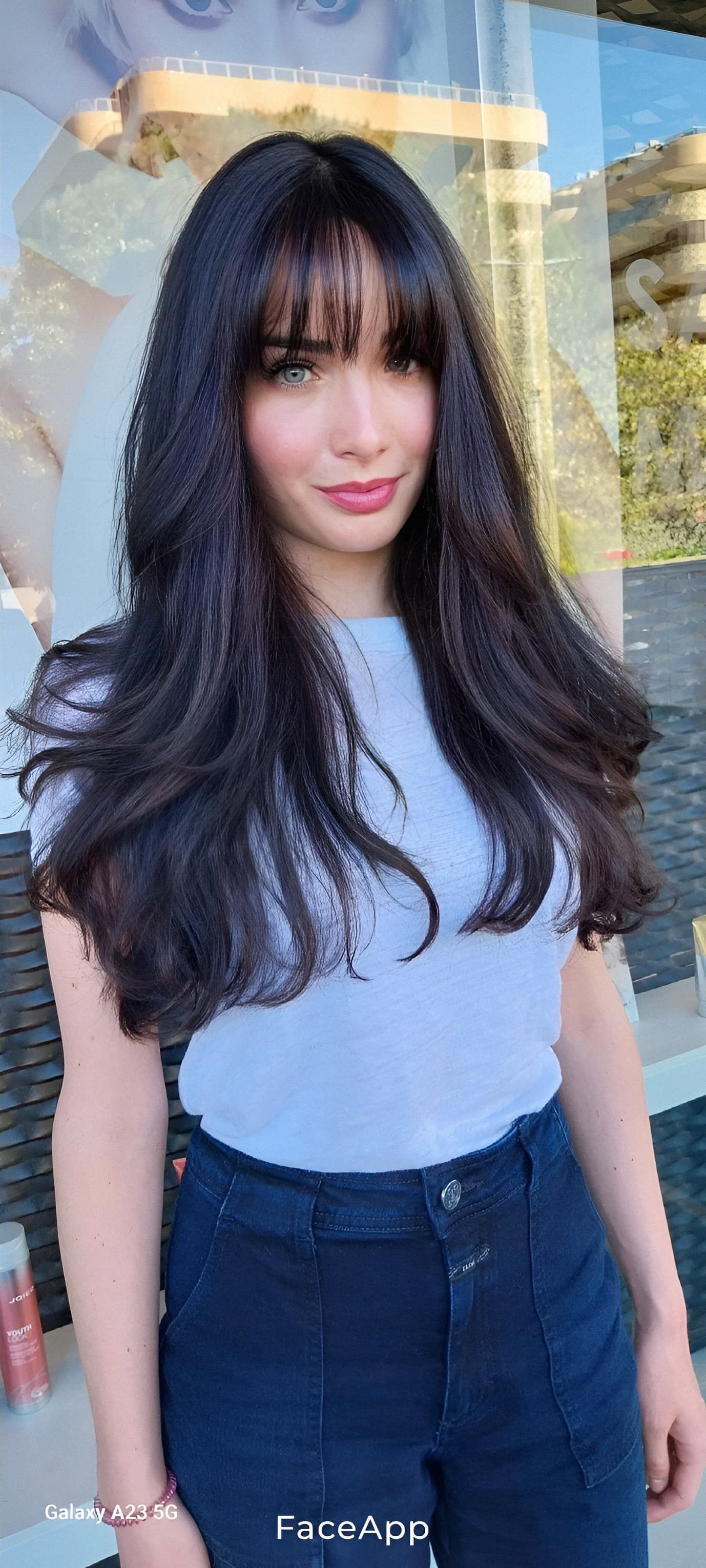Una persona con lunghi capelli scuri e ondulati e la frangia indossa una maglietta azzurra e jeans scuri davanti a una vetrina.
