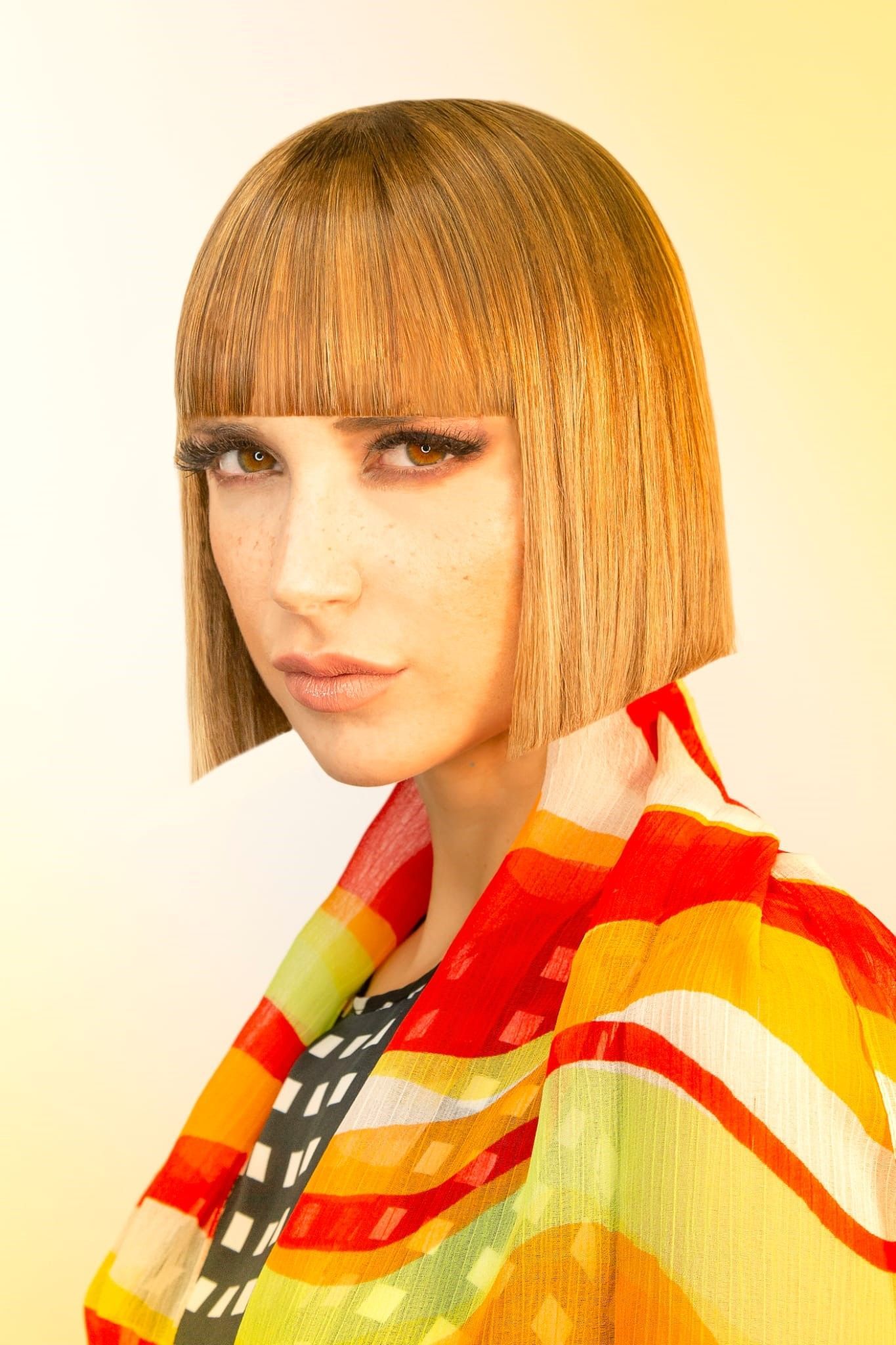 Una persona con capelli castano chiaro, mossi e tagliati a caschetto, con frangia dritta, indossa uno scialle a righe multicolori su uno sfondo giallo.
