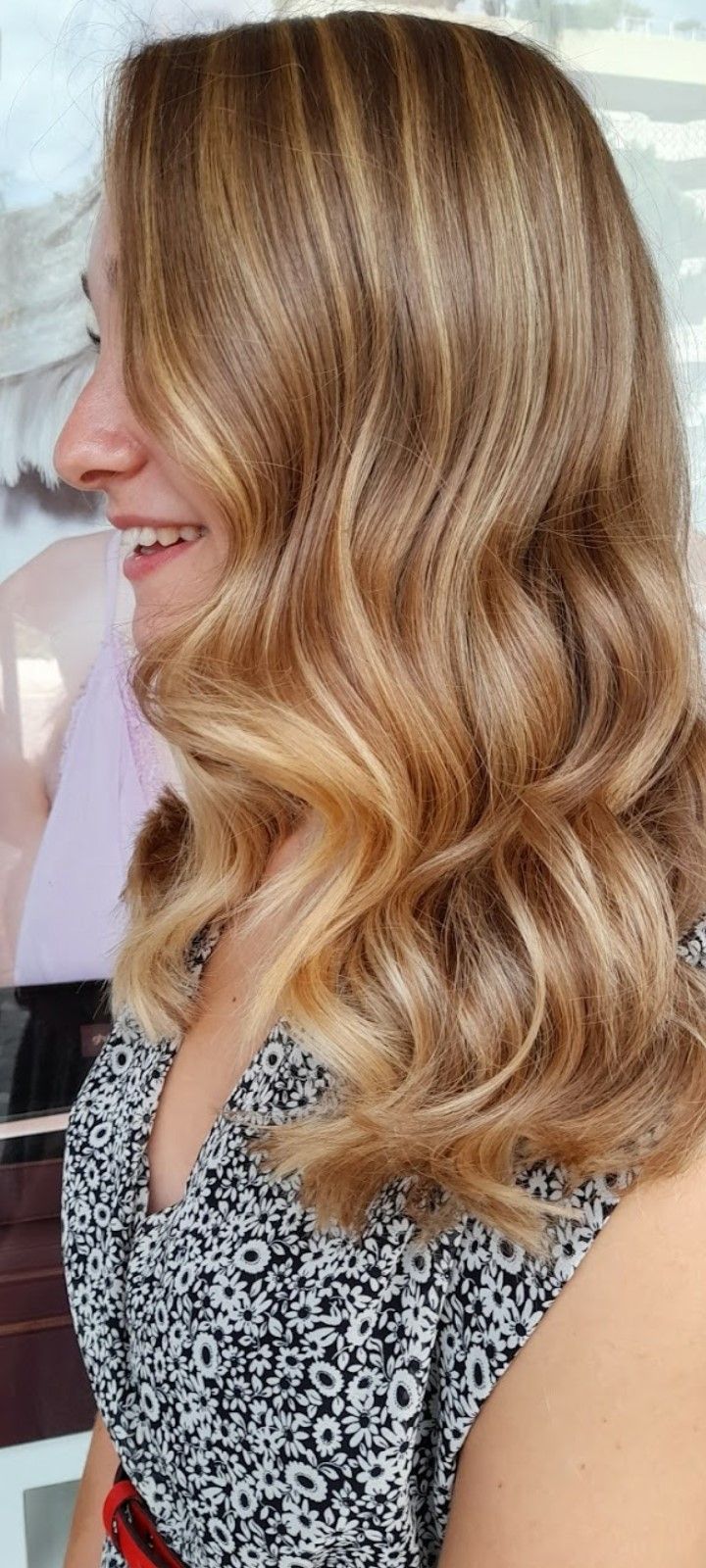 Una persona con capelli biondi ondulati e con mèches, che indossa una maglietta a fantasia bianca e nera e sorride di profilo.