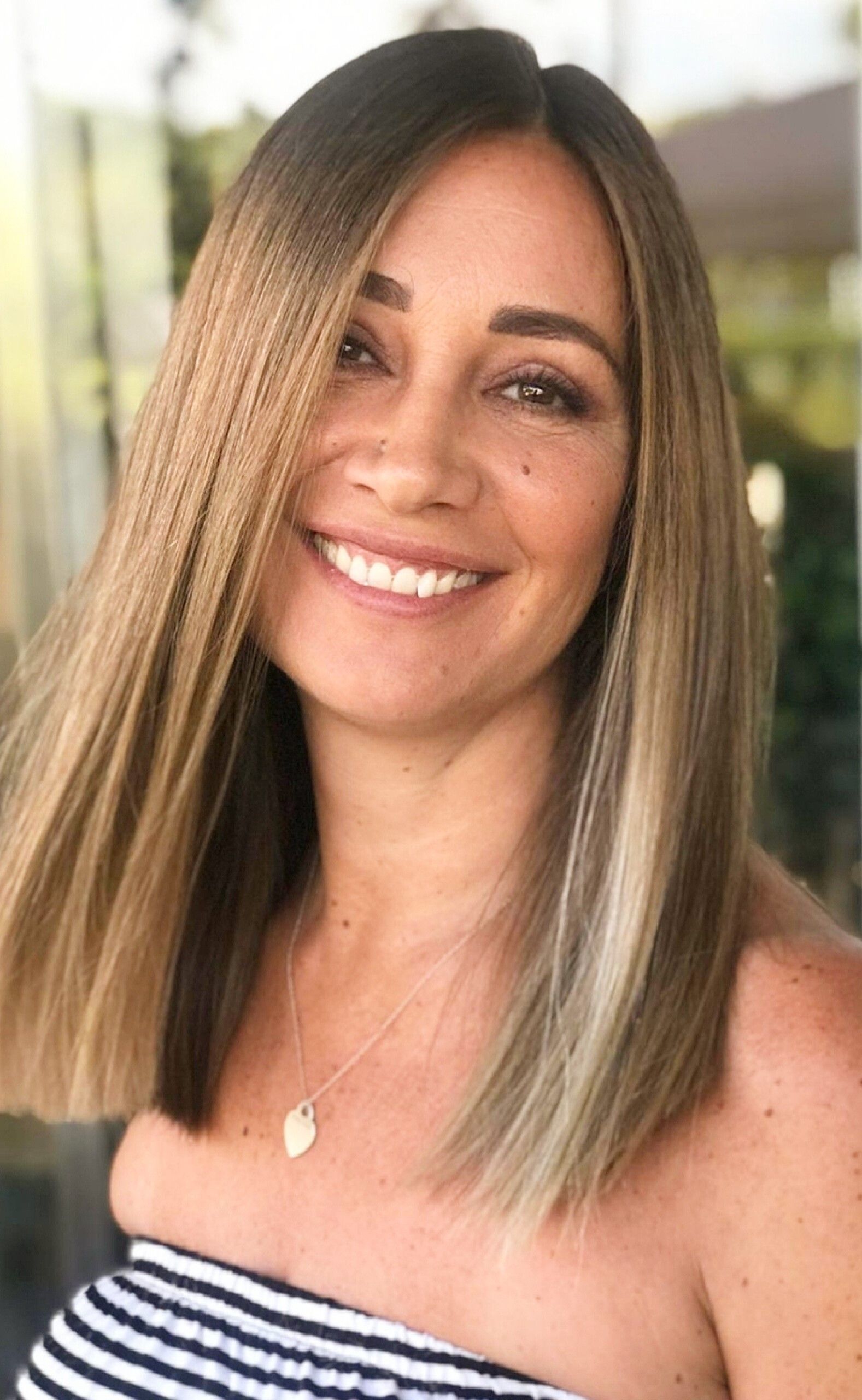 Una persona con lunghi capelli biondi con mèches sorride alla telecamera, indossando una maglietta a righe e una piccola collana con ciondolo.