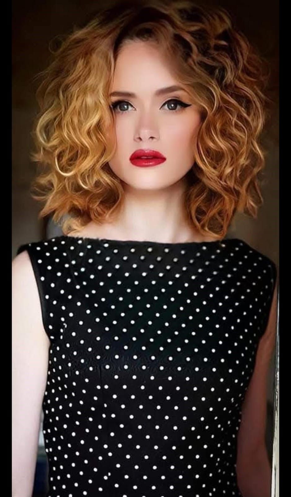 Una persona con i capelli biondi ricci lunghi fino alle spalle indossa un abito nero a pois bianchi e un rossetto rosso acceso.