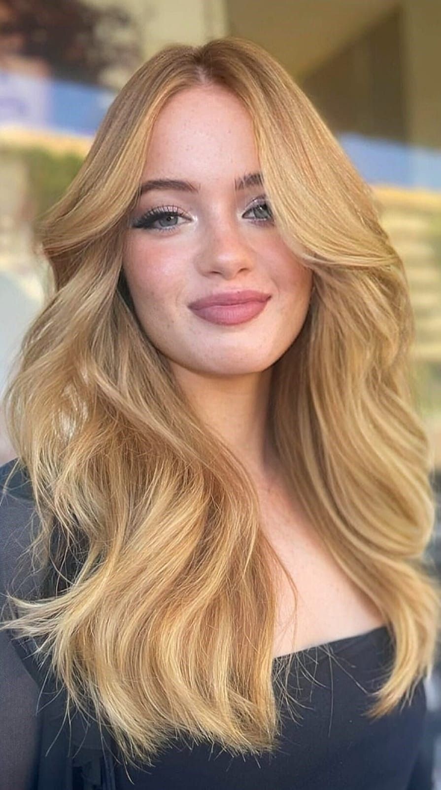 Una persona con capelli biondi ondulati di media lunghezza e un sorriso gentile, che indossa una maglietta nera.