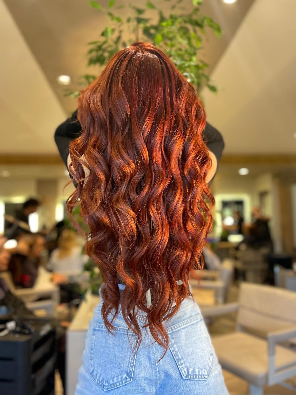 Una persona con lunghi capelli mossi di un rosso ramato intenso, ripresa di spalle, indossa una maglietta nera e jeans azzurri in un salone di bellezza.