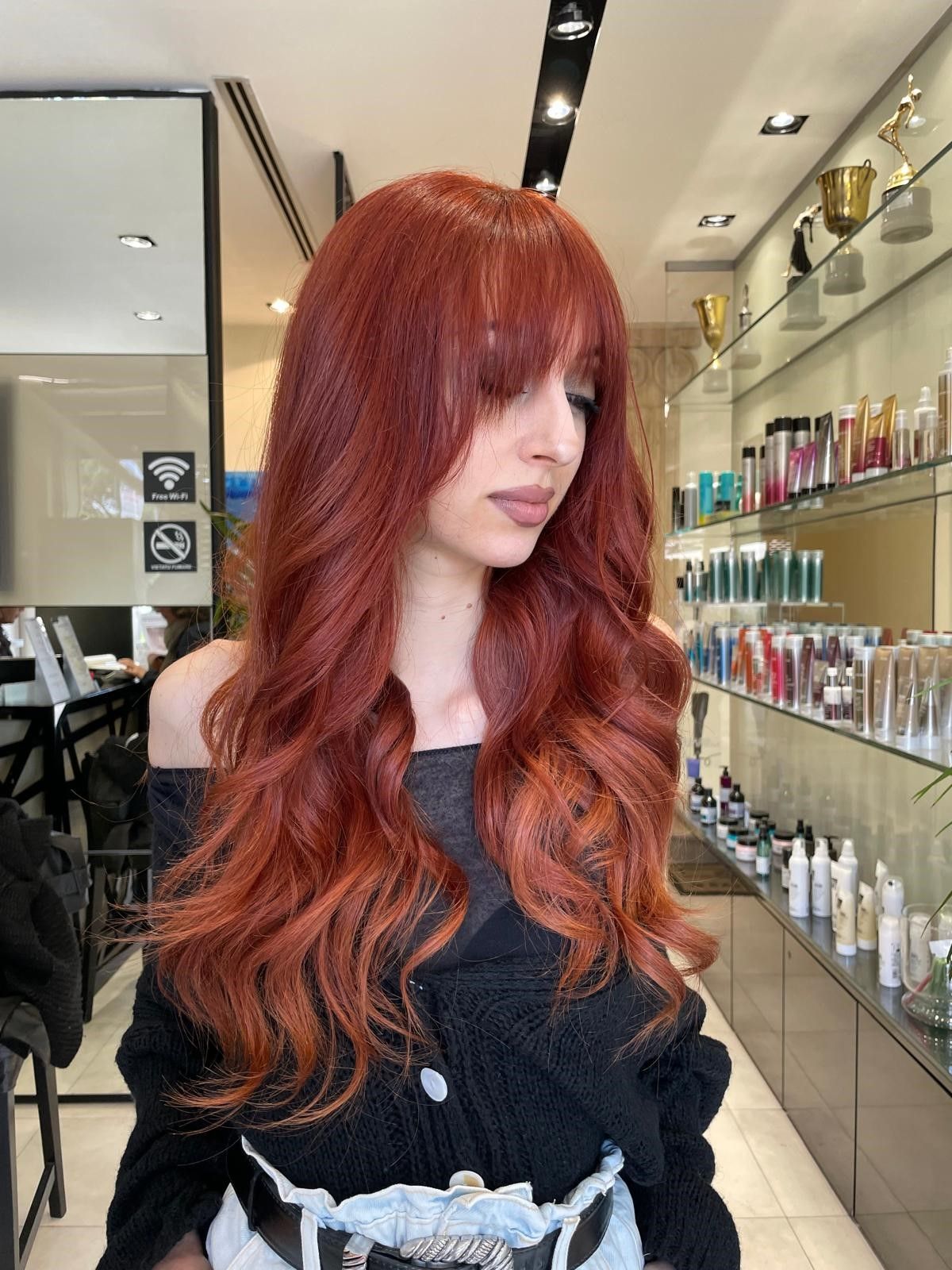 Una persona con lunghi capelli mossi color castano ramato e frangia a tendina si trova in un salone da parrucchiere pieno di prodotti per la cura dei capelli.