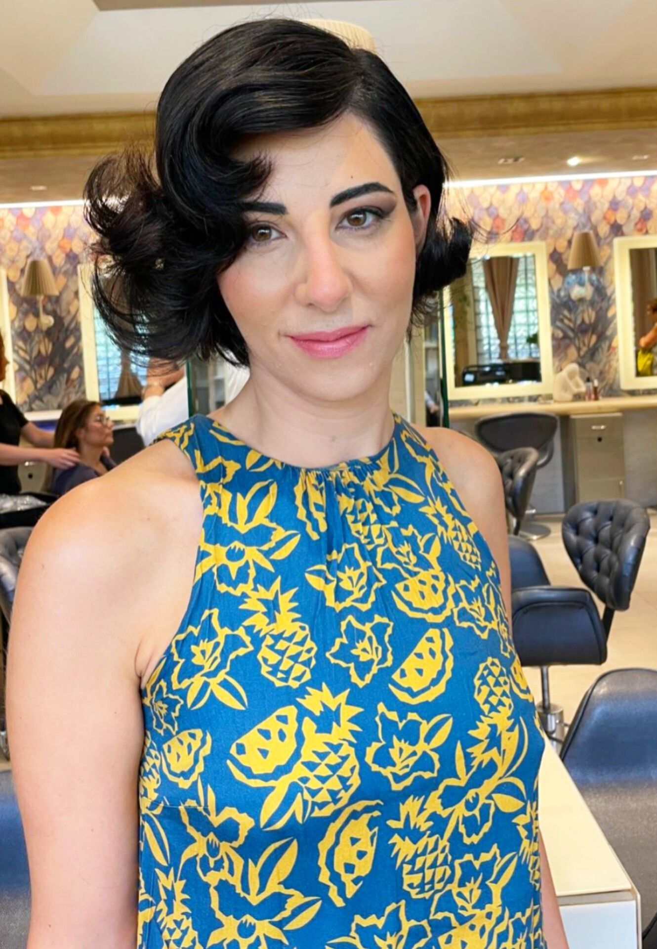 In un salone ben illuminato, una persona con i capelli corti, scuri e ricci indossa una maglietta con una fantasia di frutta blu e gialla.