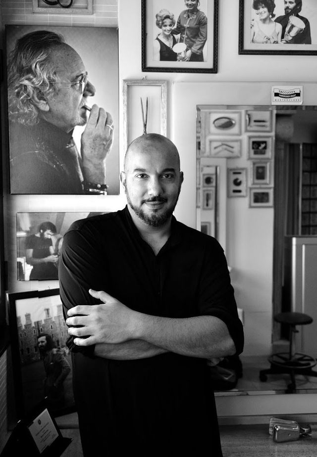 Un uomo barbuto con una camicia nera, in piedi con le braccia incrociate, in un ufficio decorato con ritratti e opere d'arte incorniciate.