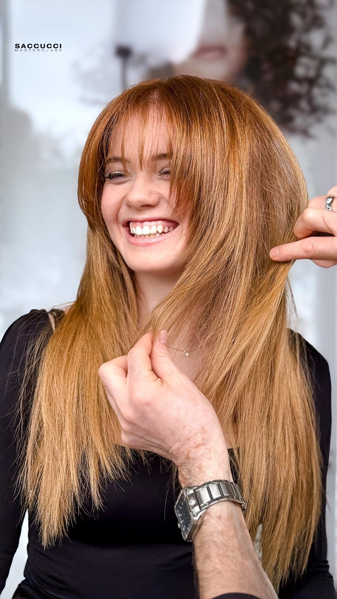 Una persona sorridente con lunghi capelli mossi color castano dorato si sta facendo acconciare i capelli da delle mani che impugnano un pettine.