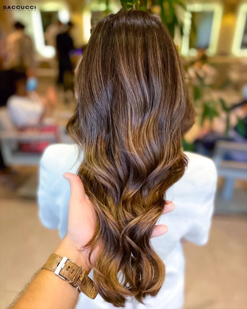 Lunghi capelli castani ondulati con riflessi caramello, mostrati in un salone da un parrucchiere che ne tiene una ciocca.