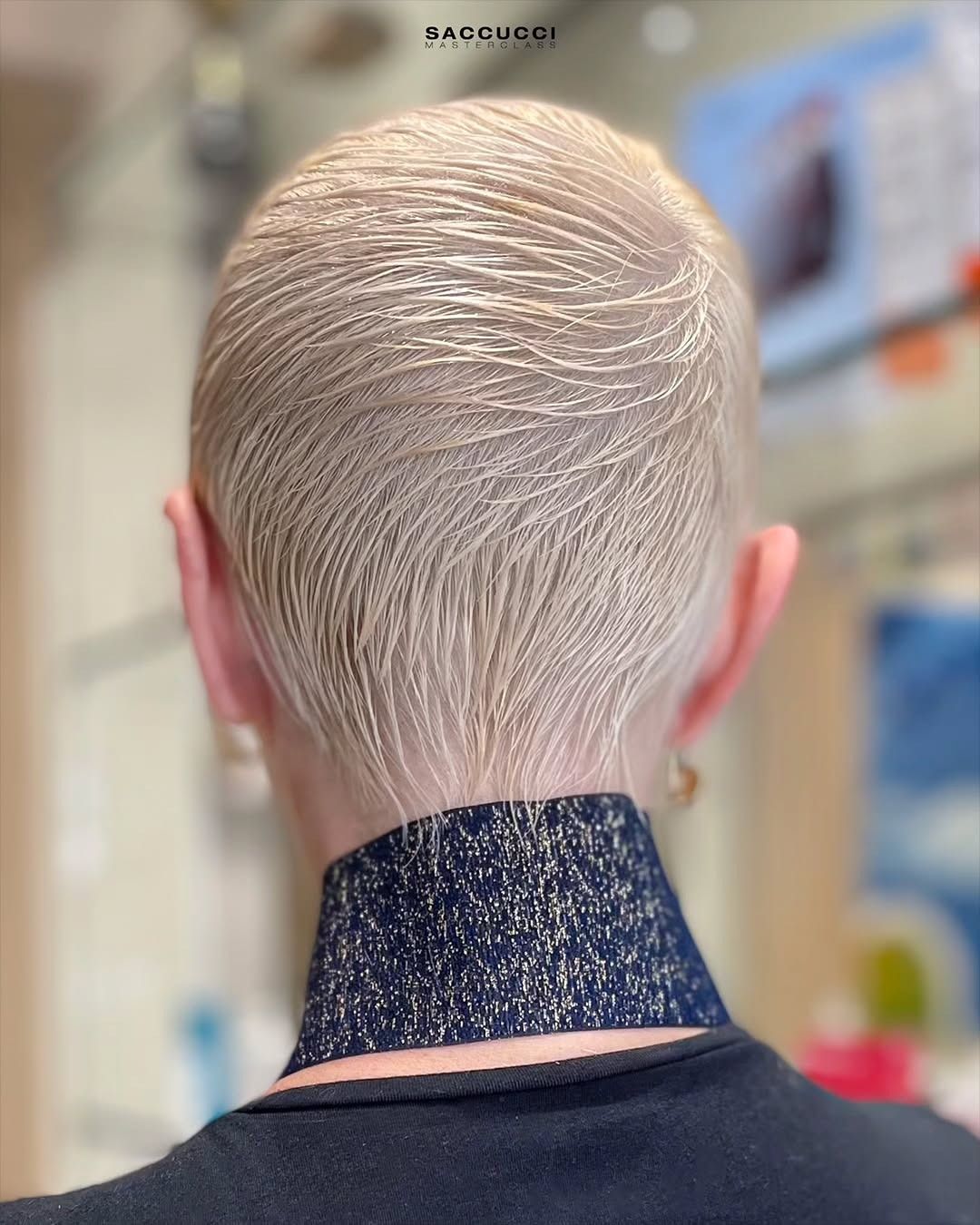 Una persona con i capelli corti, biondo platino, rasati a zero, con sottili motivi a spirale sopra un girocollo scuro e scintillante.