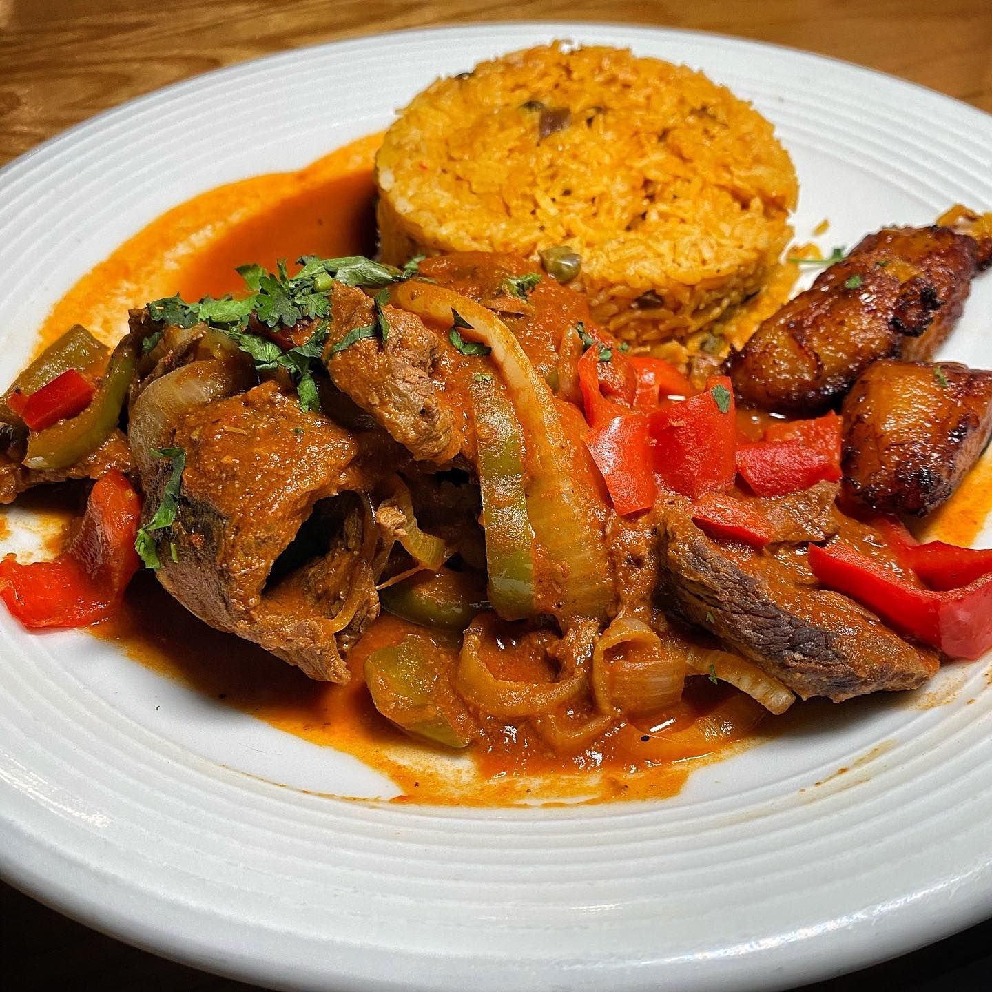 Plate of food featuring stewed meat, yellow rice, fried plantains, and peppers.