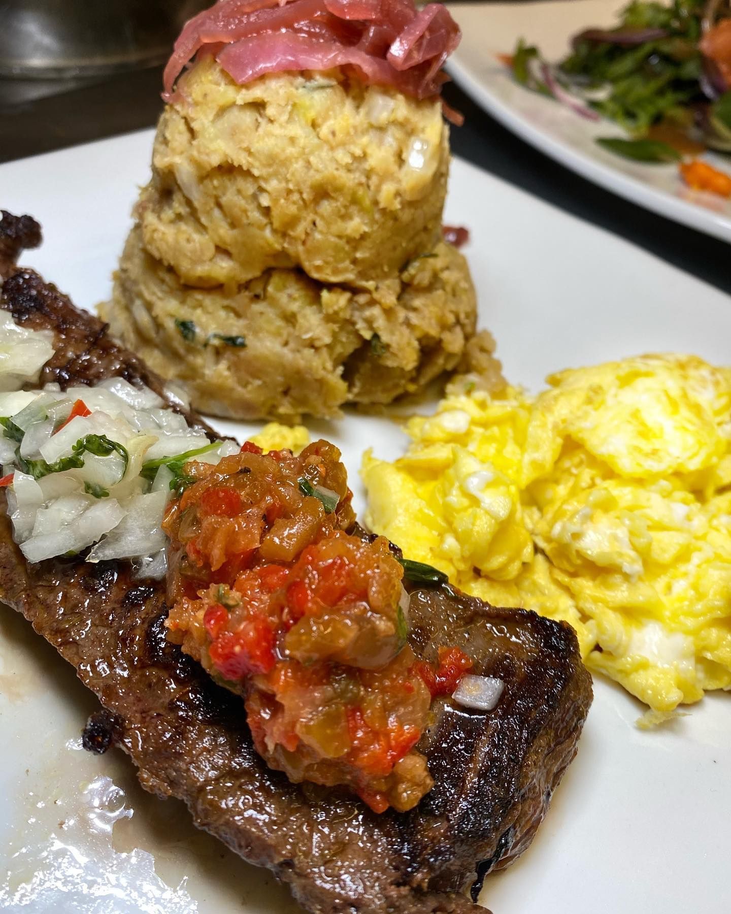 Plate of food: grilled steak with toppings, scrambled eggs, and mashed vegetable balls.