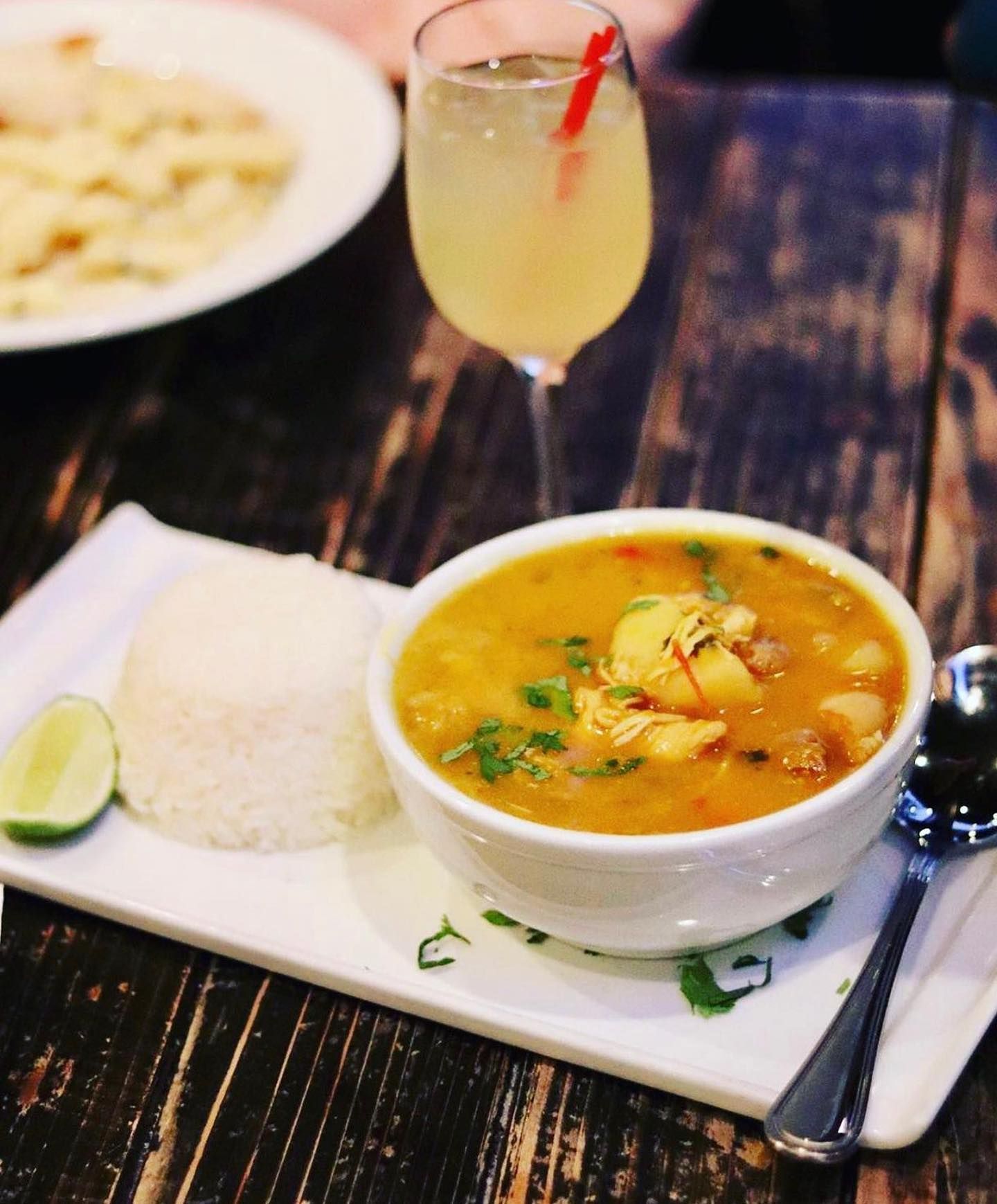 Bowl of soup with rice, lime, and a cocktail on a wooden table.