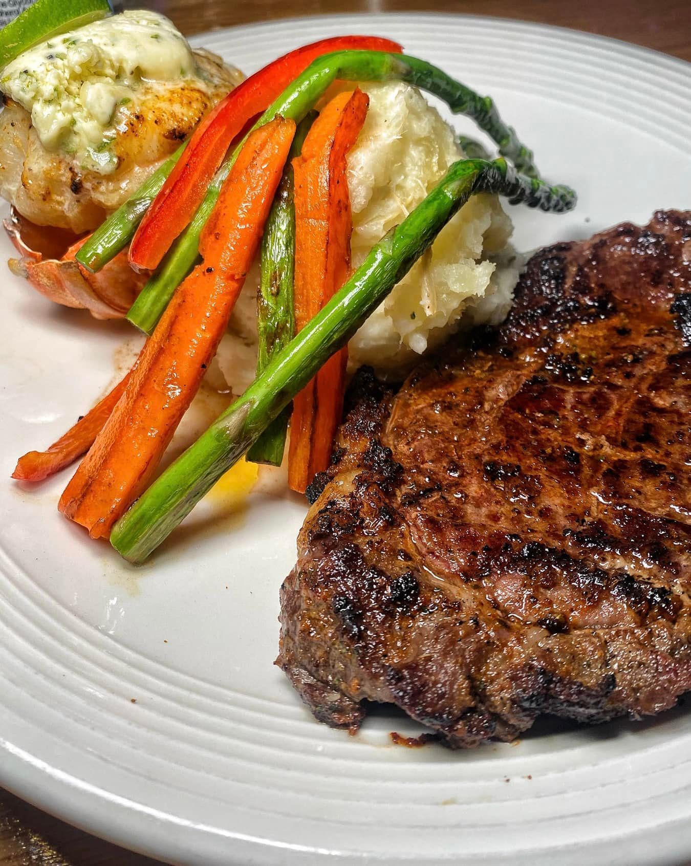 Grilled steak and lobster tail with vegetables and mashed potatoes on a white plate.