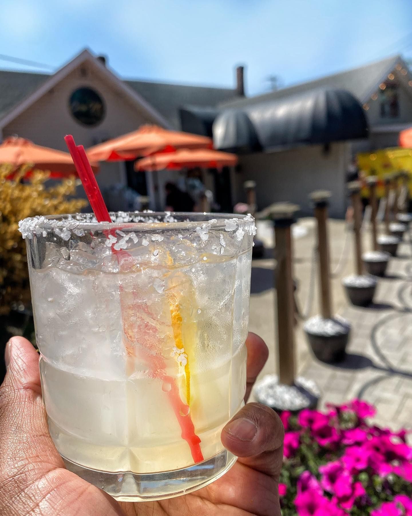 Hand holding a salt-rimmed margarita with a straw, outdoors with umbrellas and flowers in the background.
