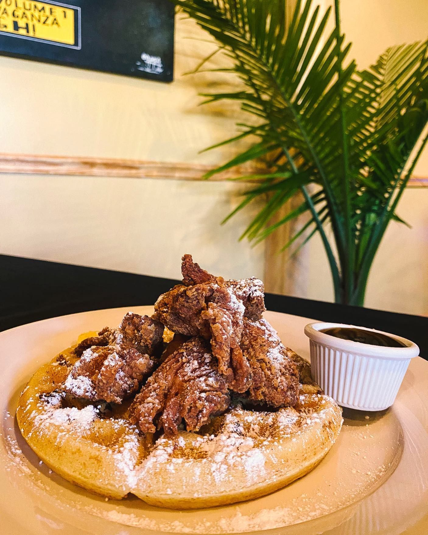 Chicken and waffles with powdered sugar and syrup, on a plate, next to a plant.