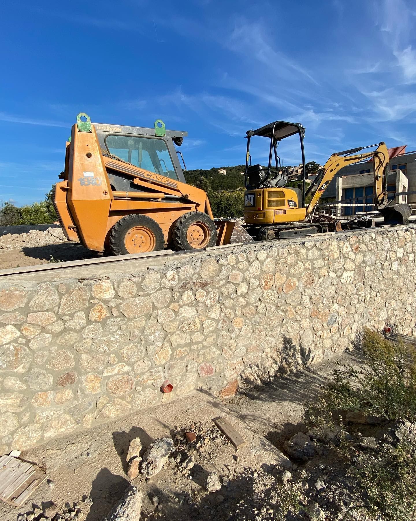 Veicoli da costruzione su un muro di contenimento in pietra sotto un cielo azzurro, tra cui una pala meccanica e un escavatore.