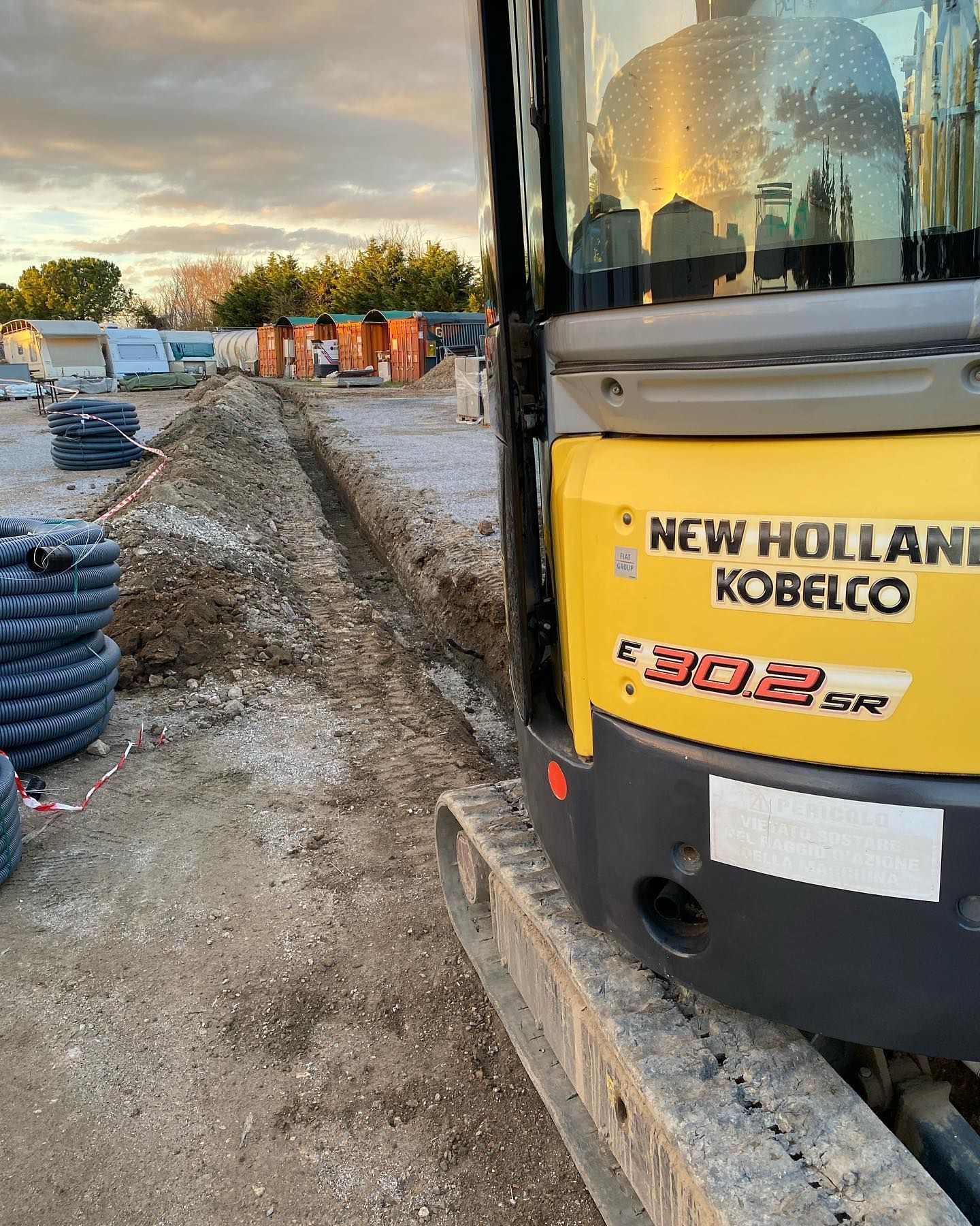Un escavatore giallo New Holland Kobelco accanto a uno scavo fangoso al tramonto.