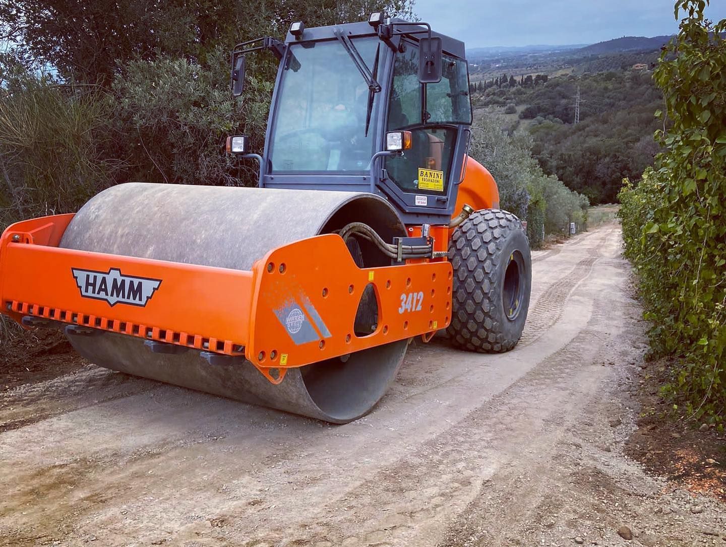 Rullo compressore arancione su un sentiero sterrato in una zona rurale boscosa.