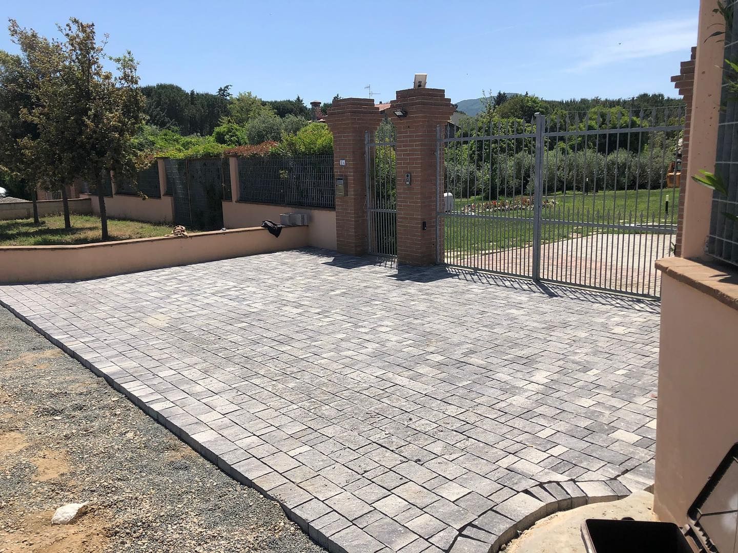 Un vialetto lastricato in pietra conduce a un cancello in mattoni e ferro, accanto a un muro color sabbia e a una zona verde.