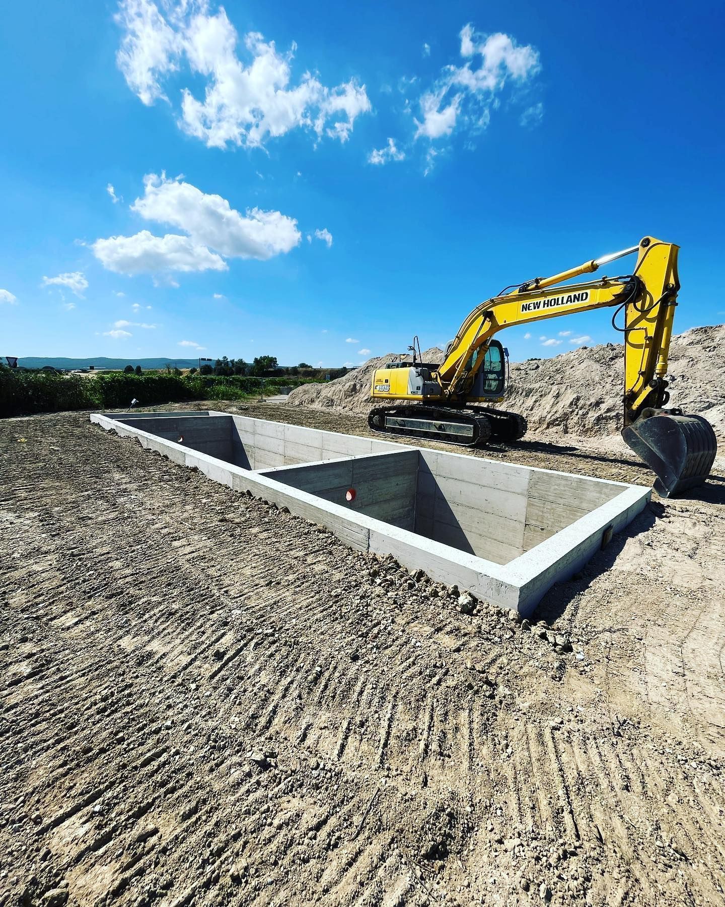 Un escavatore giallo in un cantiere edile accanto a una fondazione in cemento sotto un cielo azzurro.
