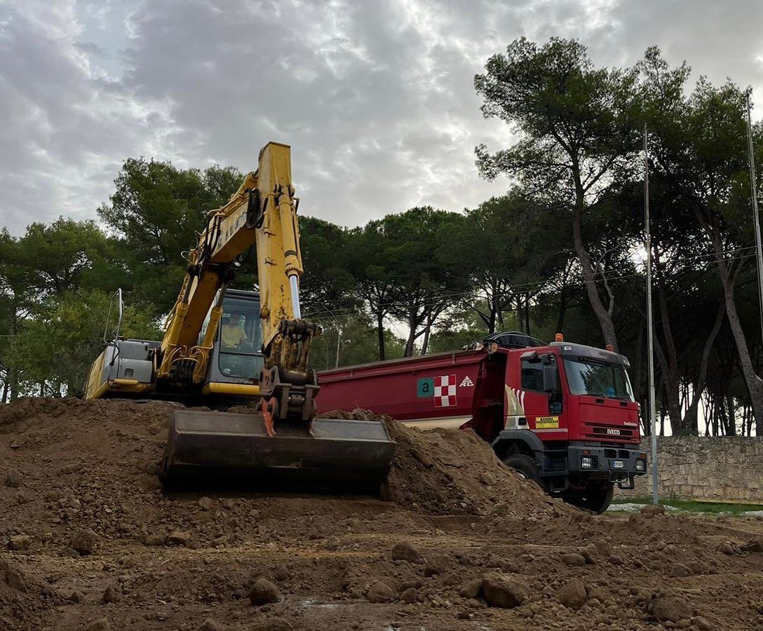 Un escavatore giallo carica della terra su un camion ribaltabile rosso in un cantiere fangoso.