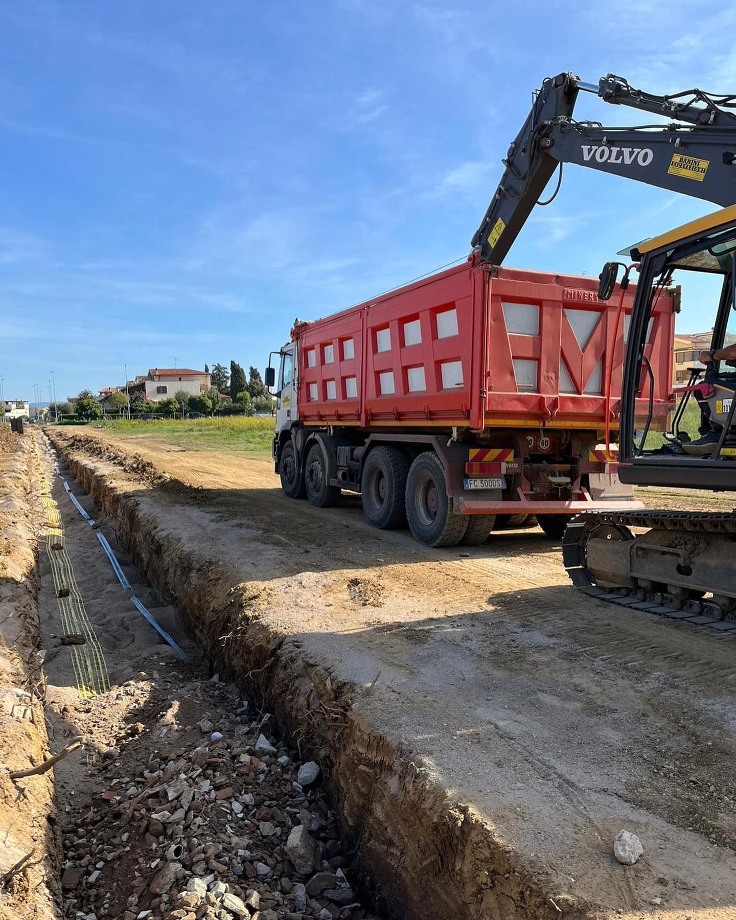 Un escavatore carica della terra su un camion ribaltabile rosso in un cantiere stradale.