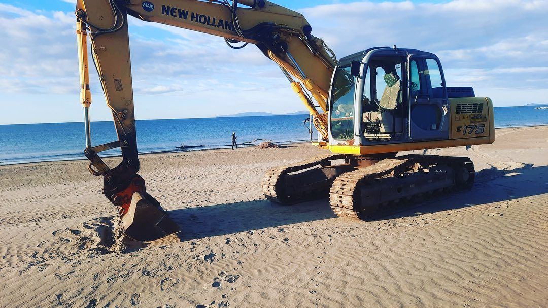 Escavatore su una spiaggia sabbiosa, intento a scavare vicino alla riva sotto un cielo azzurro e limpido.