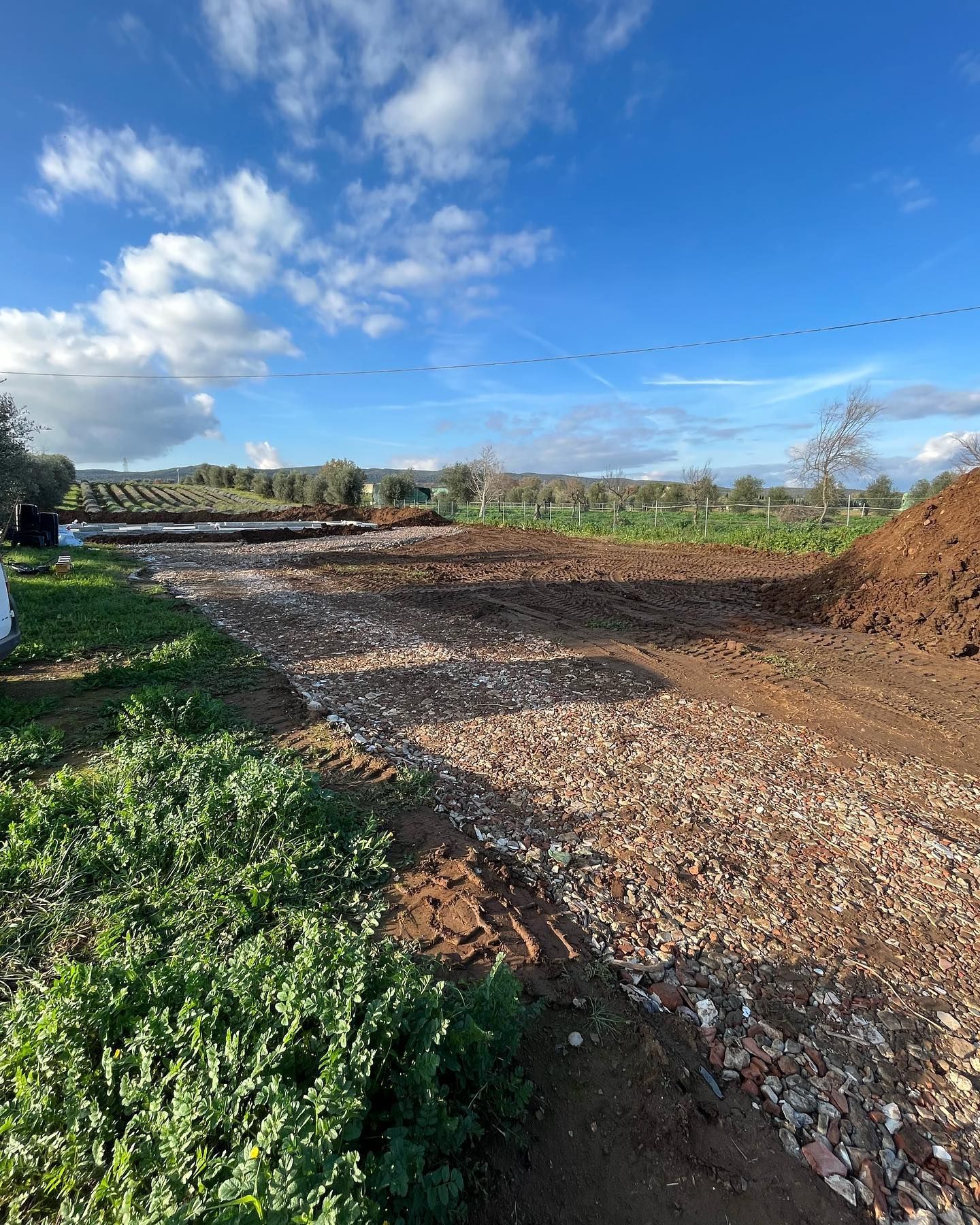 Cantiere sterrato con sentiero di ghiaia, cumuli di terra e cielo azzurro sopra un campo verde.
