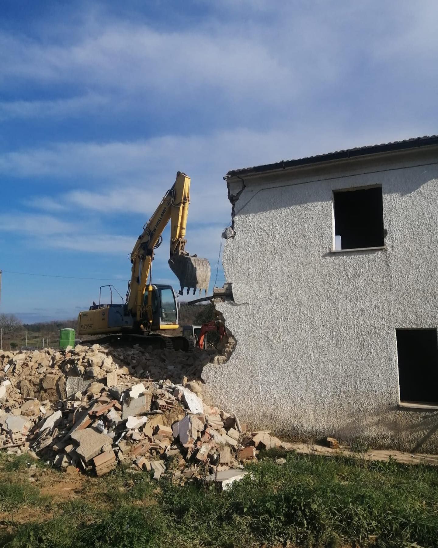 Un escavatore demolisce una casa bianca accanto a un cumulo di macerie sotto un cielo azzurro.