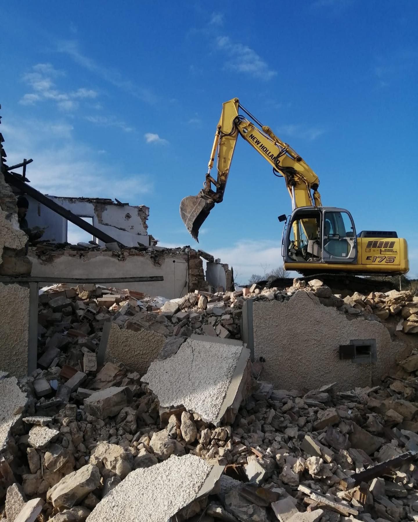Un escavatore giallo demolisce un edificio ricoperto di macerie sotto un cielo azzurro.