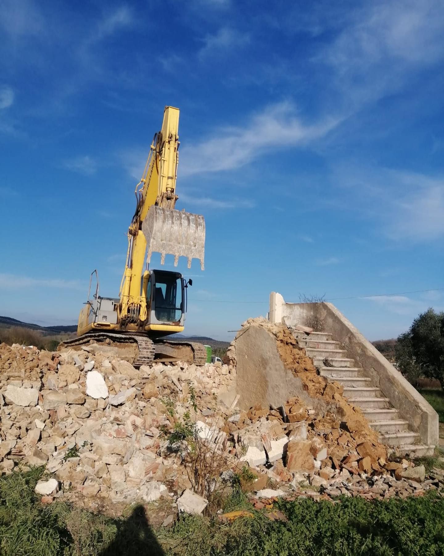 Escavatore giallo tra le macerie accanto a una scala di cemento rotta sotto un cielo azzurro.