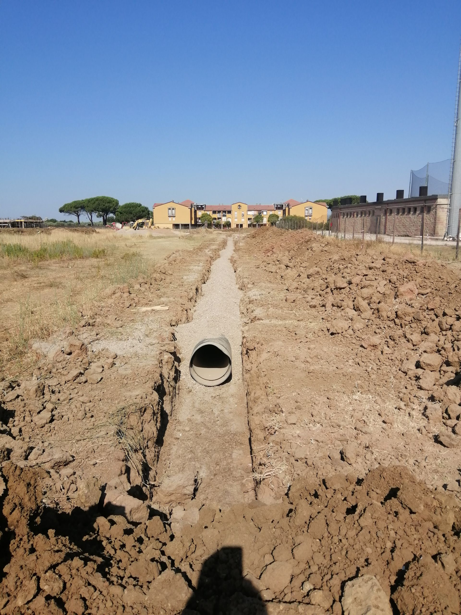 Tubo di drenaggio in cemento in uno scavo di terra, con case sullo sfondo, sotto un cielo azzurro e limpido.