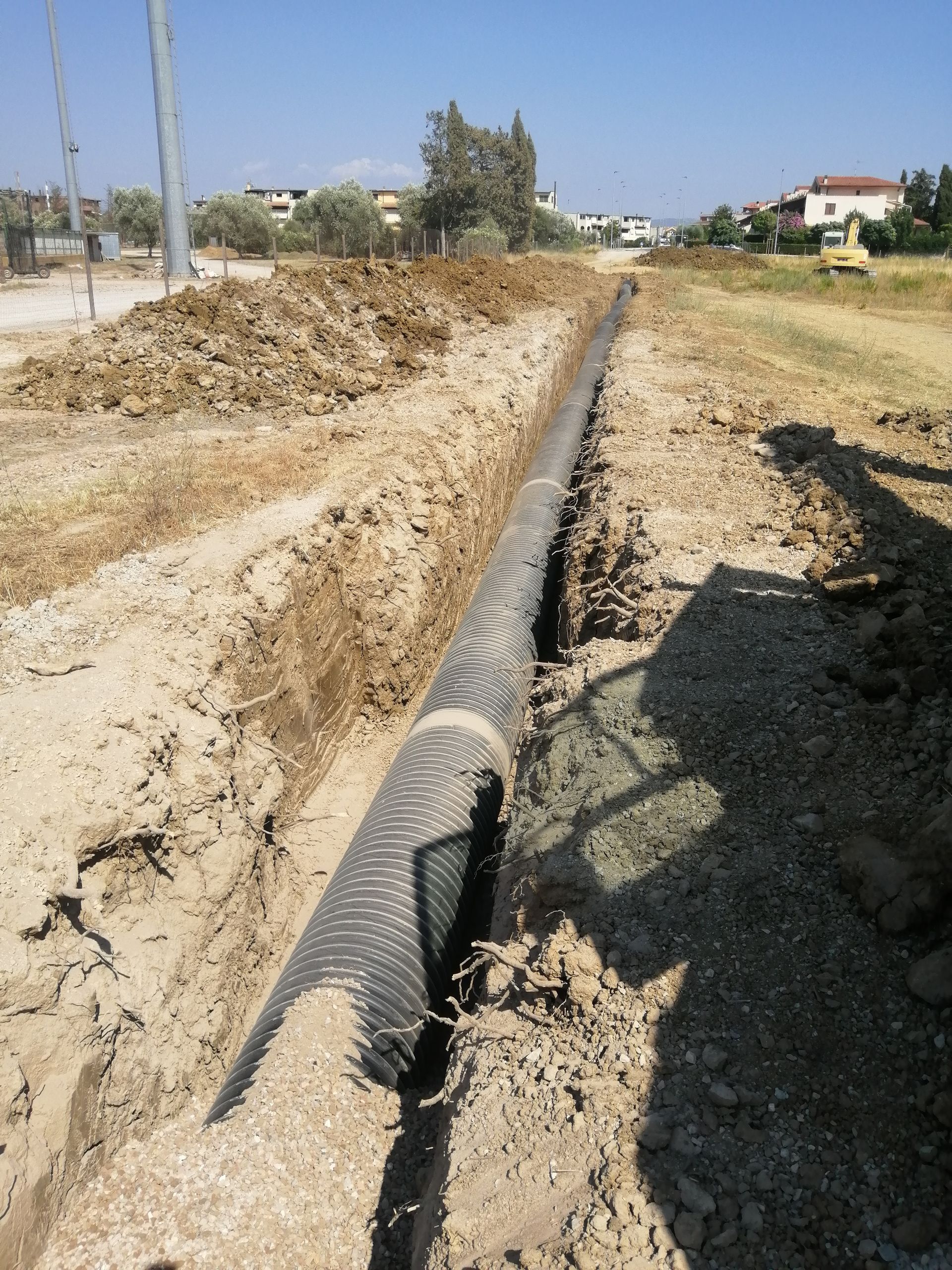 Una lunga conduttura posata in una trincea di terra a lato di una strada rurale, con campi e case sullo sfondo.