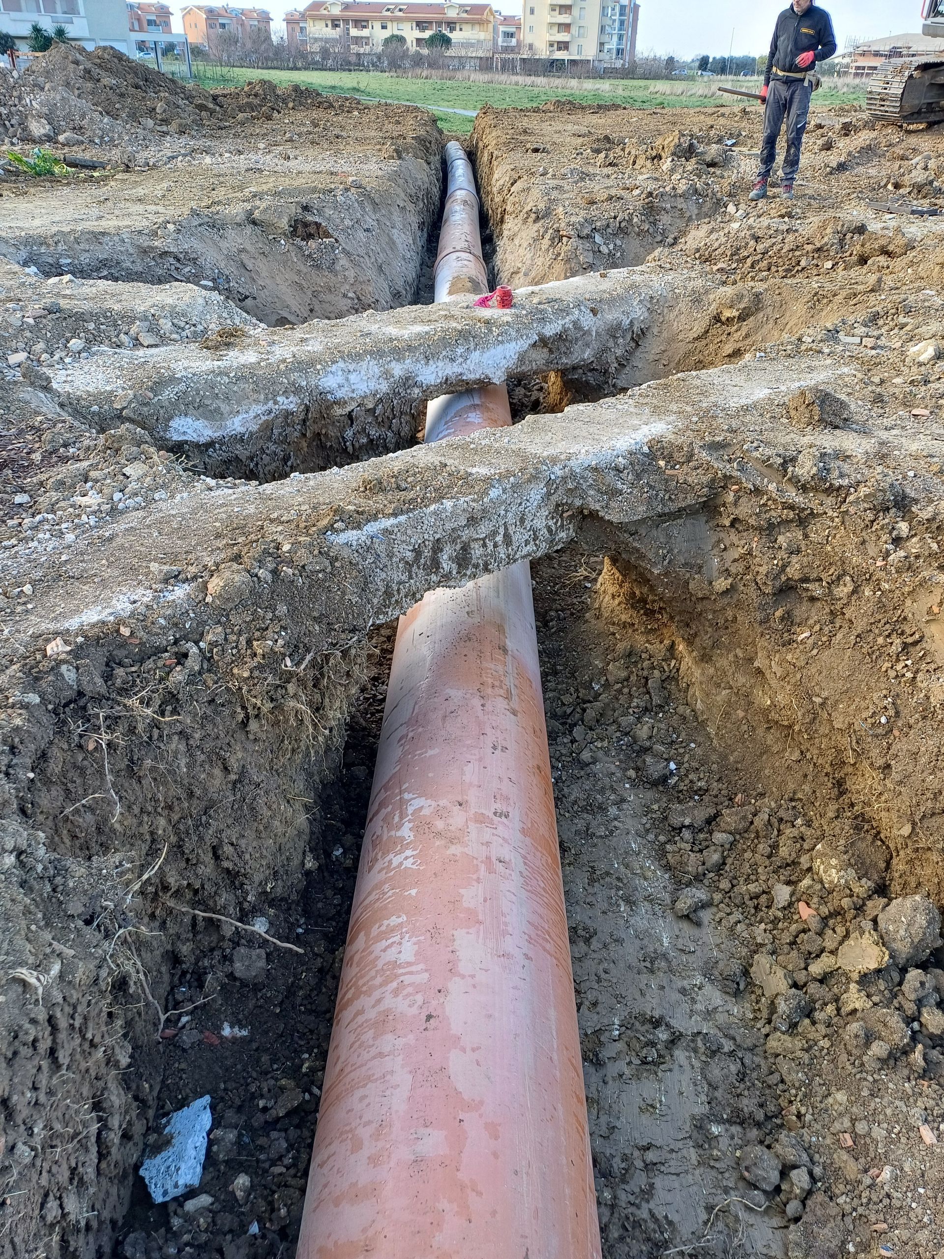Una grande conduttura posata in una trincea con riempimento di ghiaia e un operaio in piedi nelle vicinanze all'aperto.