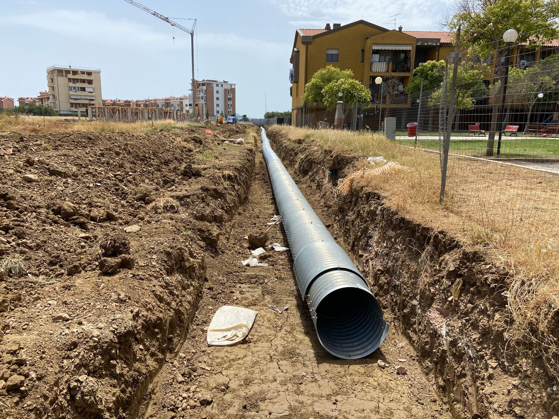 Un grosso tubo nero è stato posato in una trincea in un cantiere edile, con cumuli di terra ed edifici sullo sfondo.
