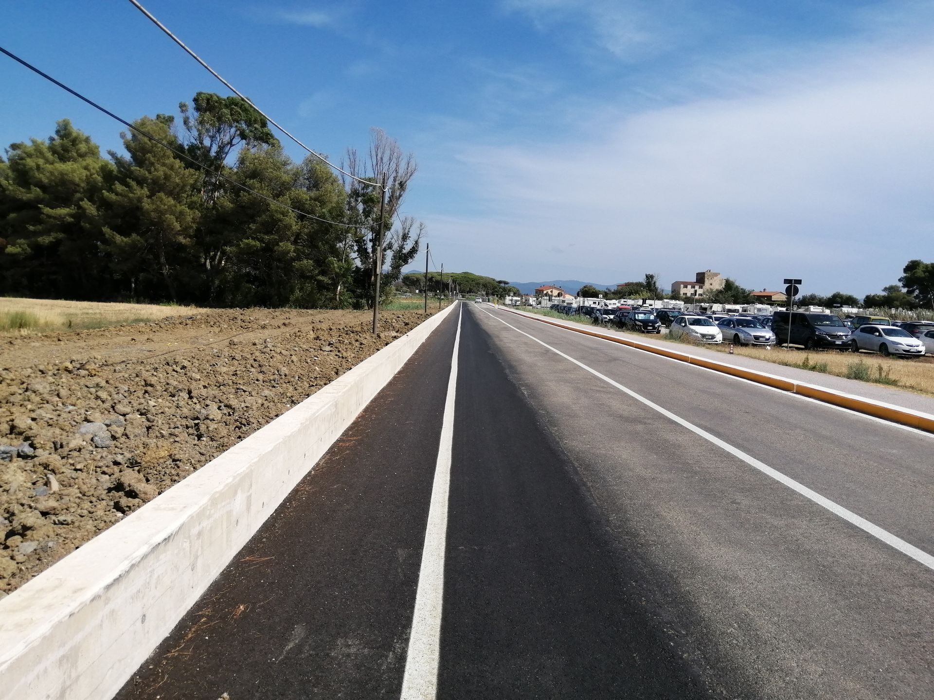 Una banchina autostradale deserta, accanto a un terrapieno erboso, con auto parcheggiate e il cielo azzurro in lontananza.