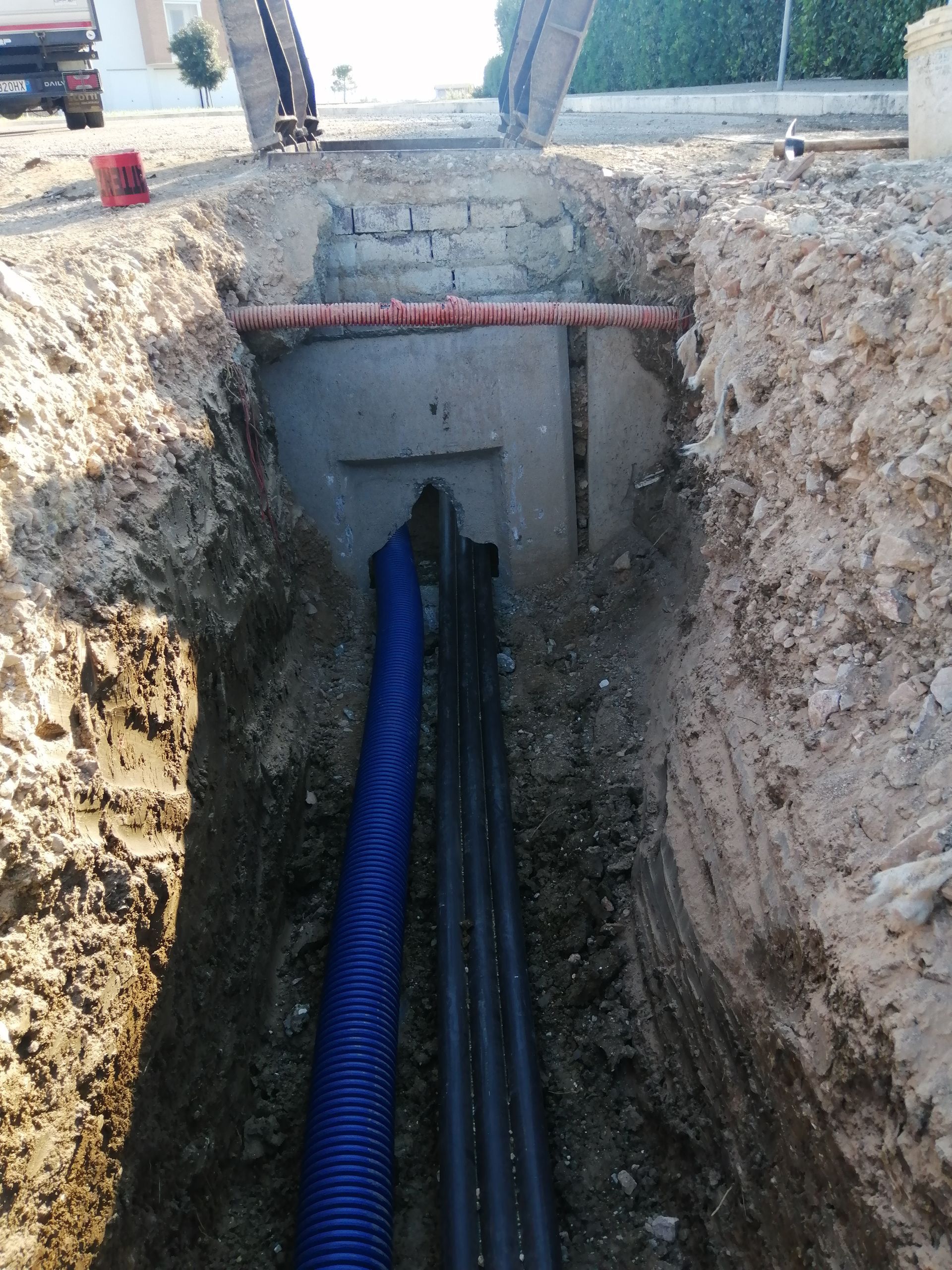Trincea di servizio con tubi di protezione blu che confluiscono in un'apertura in cemento in un cantiere.