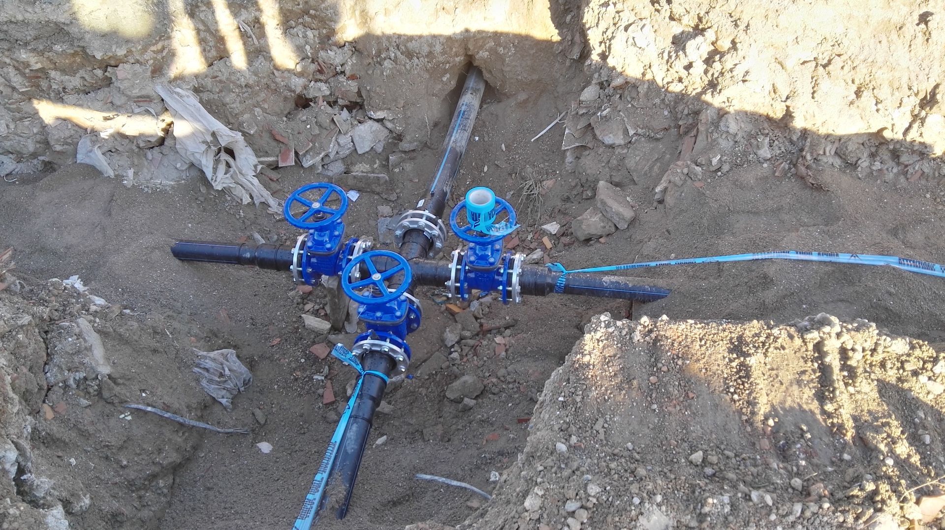Gruppo di valvole di servizio blu in una trincea di terra con tubi e terreno esposto intorno.