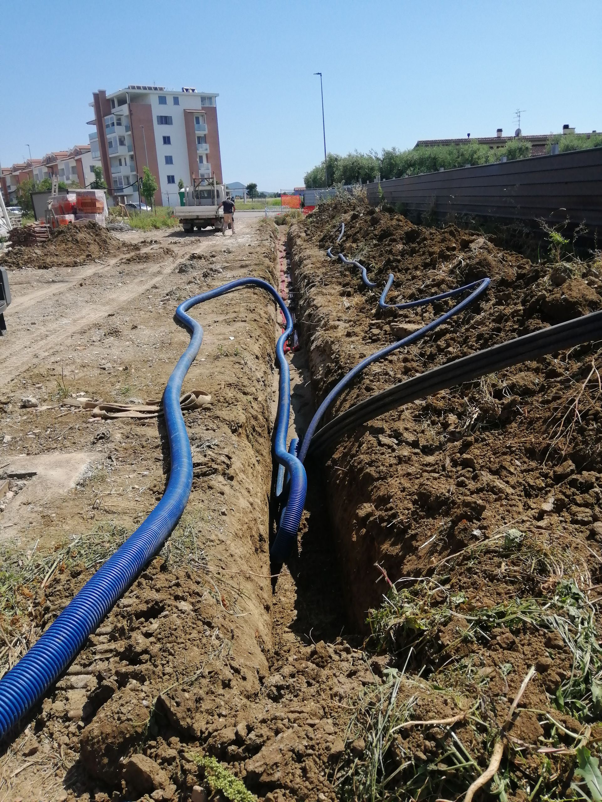 Tubi blu attraversano una trincea di terra in un cantiere edile, con edifici sullo sfondo.
