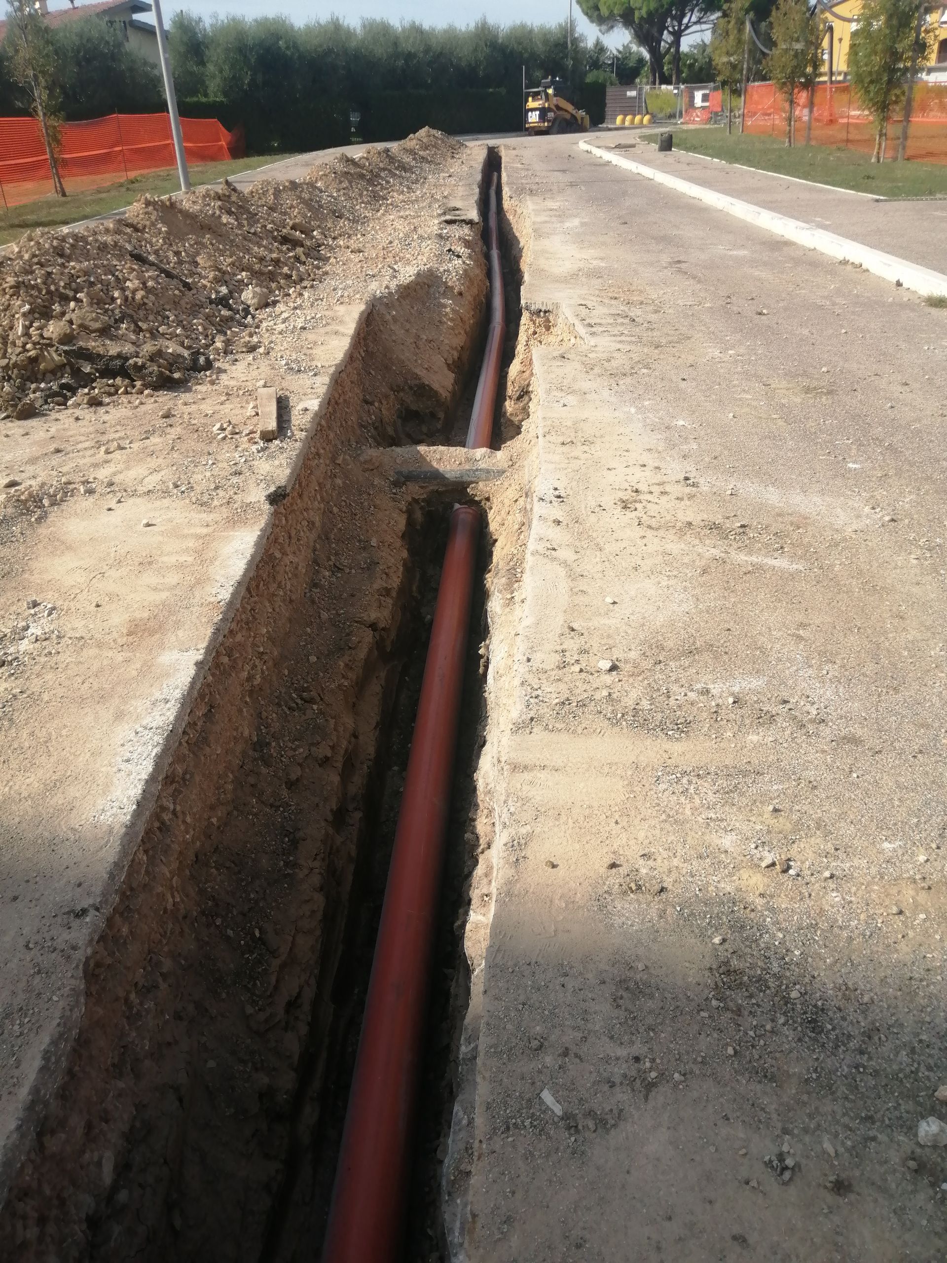 Trincea a cielo aperto con tubi rossi esposti lungo un cantiere stradale in ghiaia.