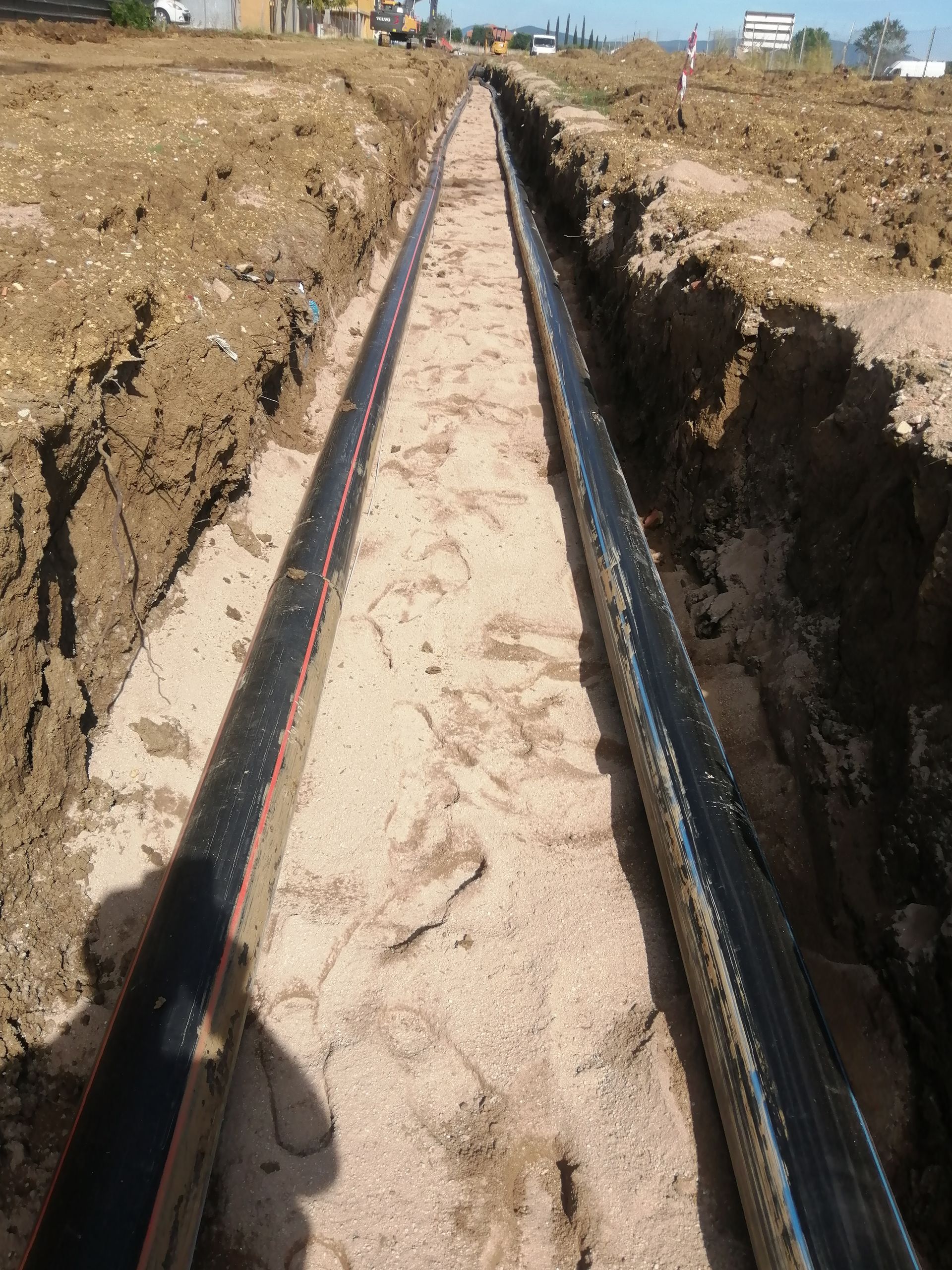 Lunga trincea scavata in terreno sabbioso e rivestita su entrambi i lati con tubi neri in un cantiere edile.