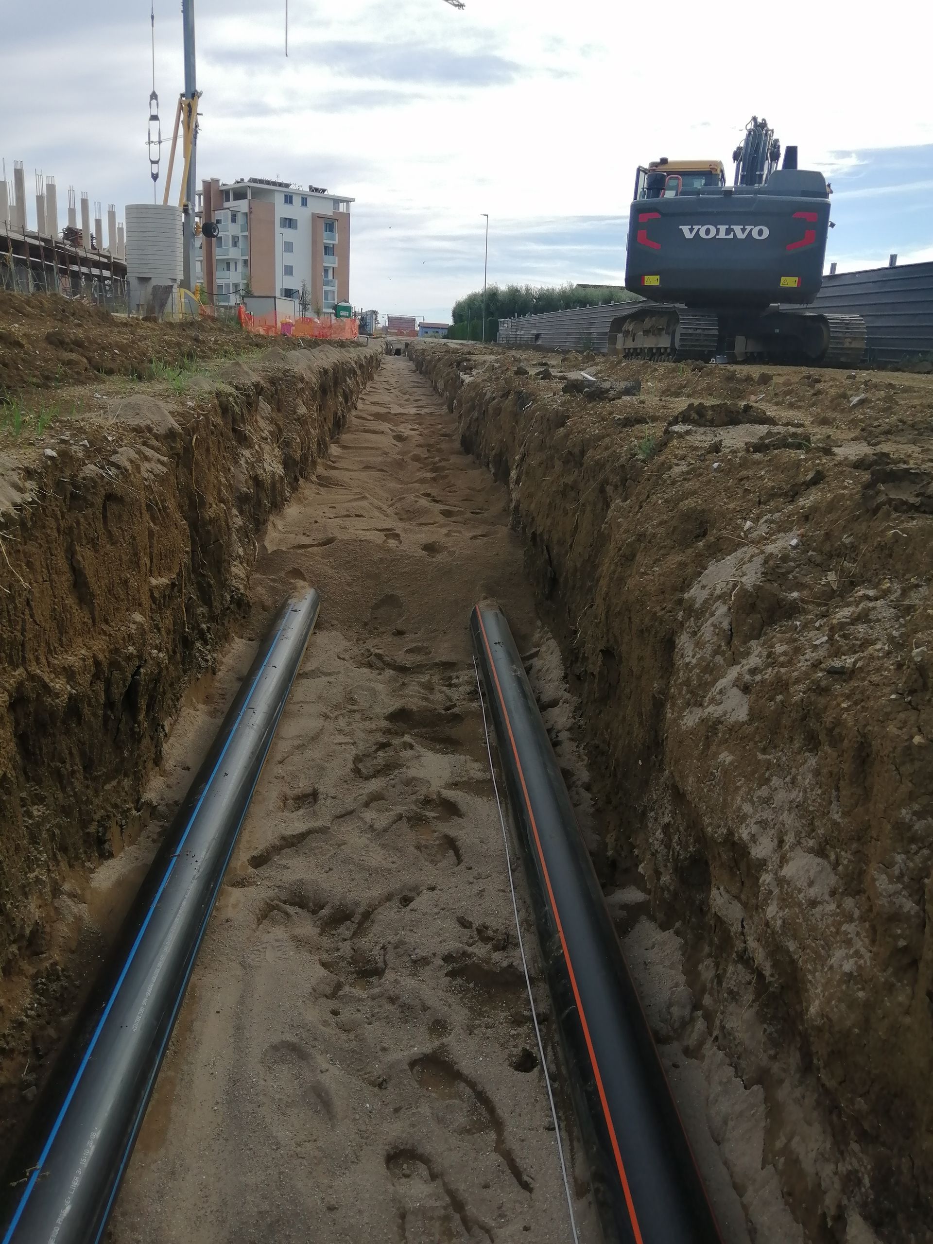 Scavo con tubature a vista in un cantiere, con un grande escavatore Volvo e degli edifici sullo sfondo.