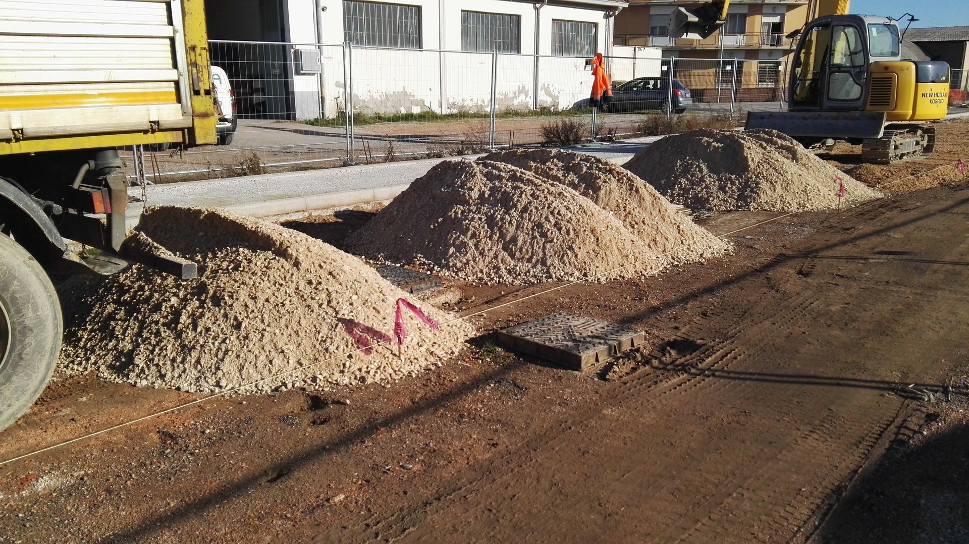 Cantiere stradale con cumuli di ghiaia, camion giallo ed escavatore a lato di una strada sterrata.