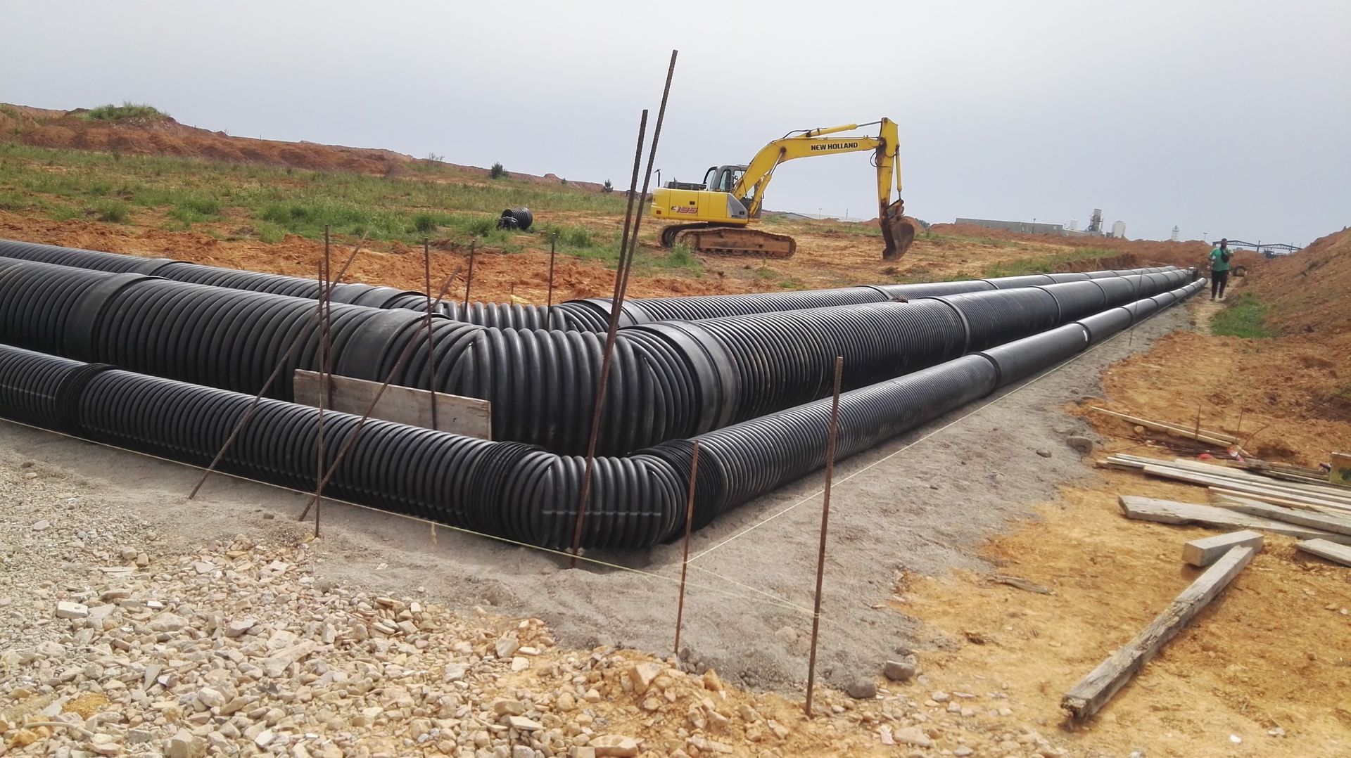 Cantiere edile con tubi di drenaggio neri, fossato di ghiaia ed escavatore sullo sfondo.