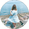 A woman in a blue dress is standing on a rocky beach looking at the ocean.